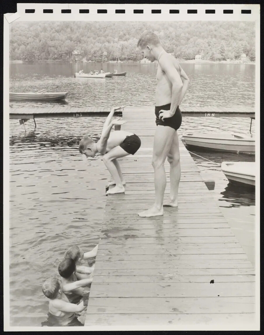 A black and white photograph of a group of boys swimming near a dock in a body of water. One boy prepares to jump from the dock, while a man standing on the dock watches on. Three boys wait in a line in the water to the left of the dock and all look at the boy about to jump. There are a couple of empty canoes that are tied to the dock.