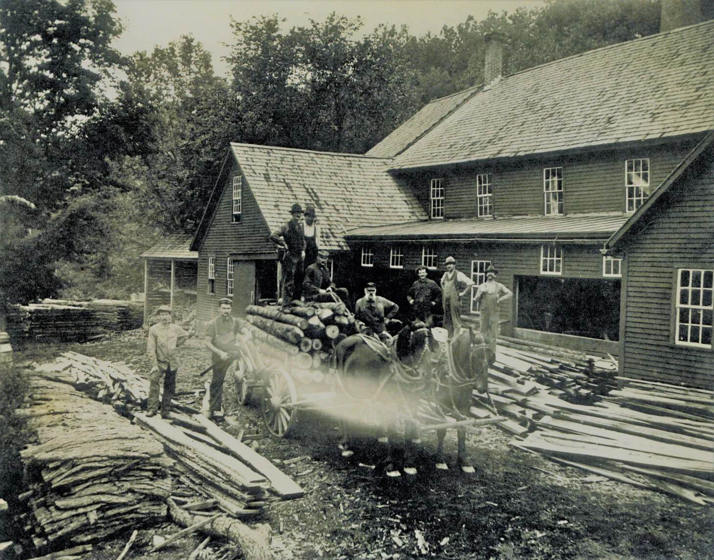 A black and white photograph of a group of men standing among piles of wood in front of a large house. Some of the men stand on top of a wagon pulled by two horses. The wagon is filled with cut logs.