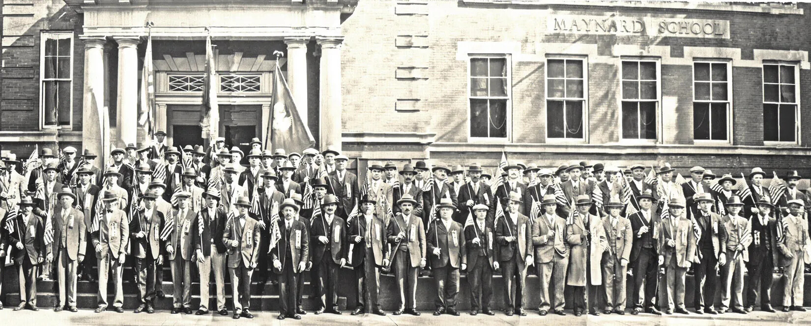 A black and white photograph of a large group of men standing on a set of stairs in front of a large building. They all look directly at the camera and hold small American flags. There is a sign on the building, which reads "MAYNARD SCHOOL."