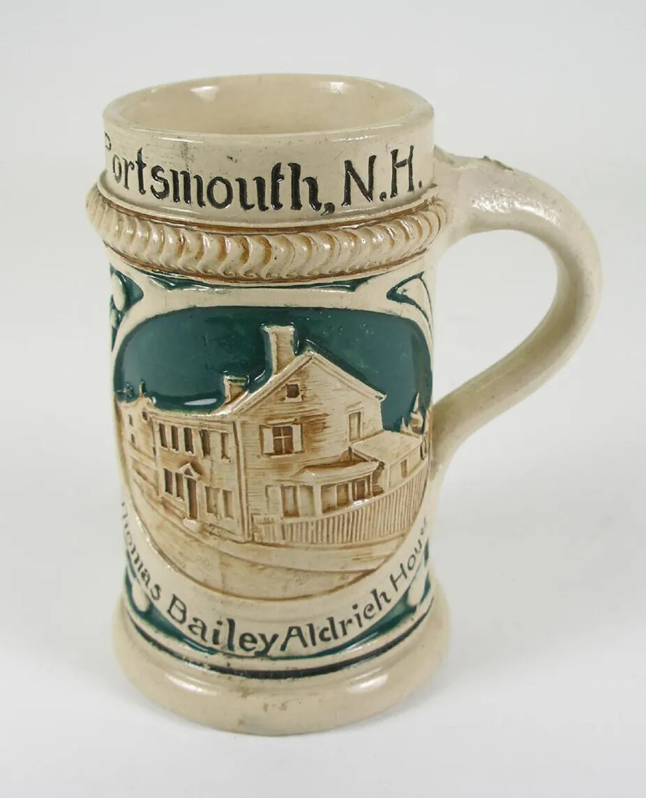 A tall drinking glass shaped like a cylinder. The glass and its handle are white. The center of the glass has a blue background and a white raised engraving of a two-storied house and fence placed along a road. Curved beneath the house are the words "Thomas Bailey Aldrich House" painted on in black paint. Engraved along the rim of the glass are the words "Portsmouth, N.H." colored in black paint. Below these is a raised, braided ribbon.