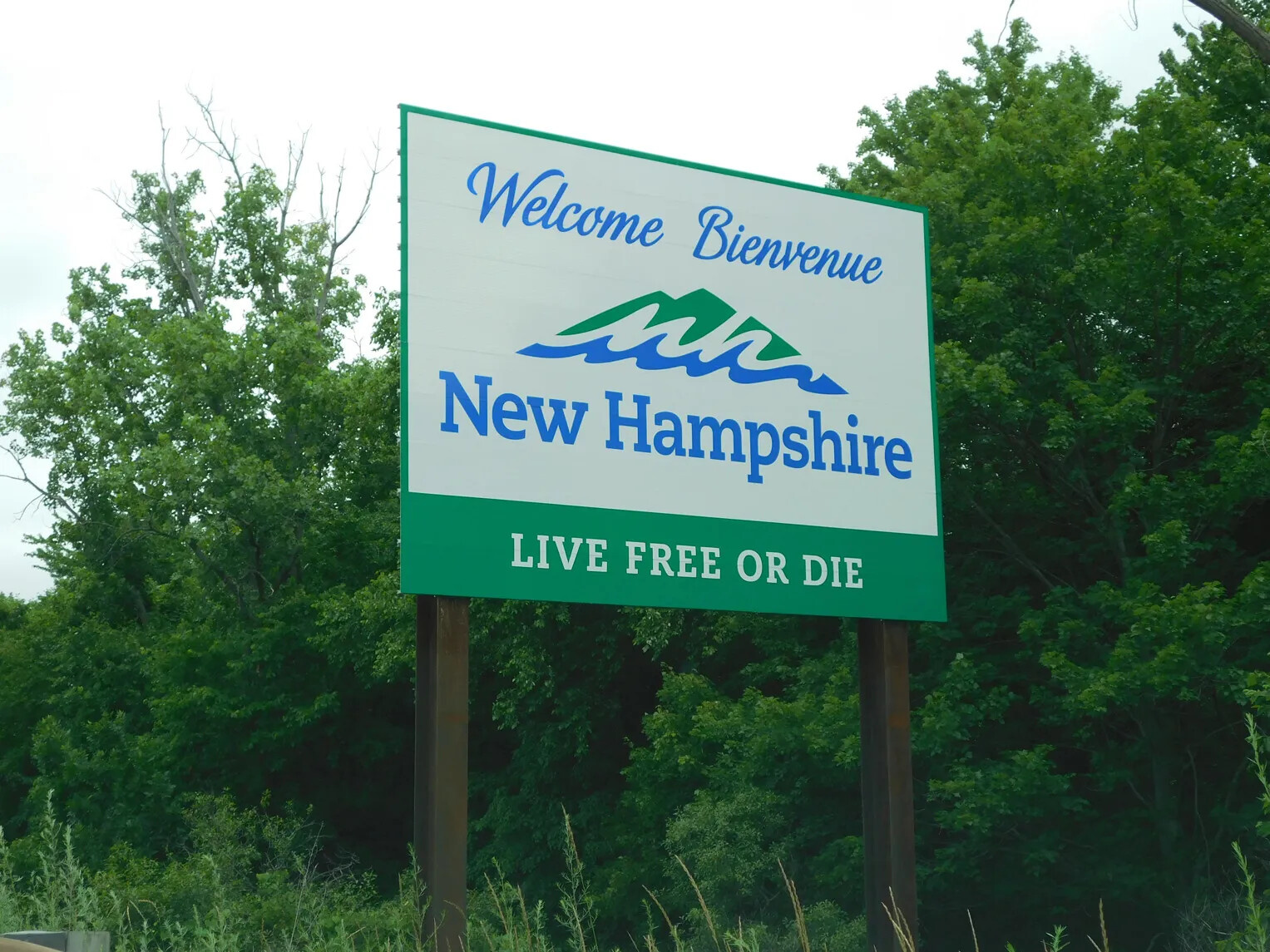 A colored photograph of a large white, green, and blue sign in front of a forested area. The sign reads: "Welcome Bienvenue/nh/New Hampshire/LIVE FREE OR DIE."