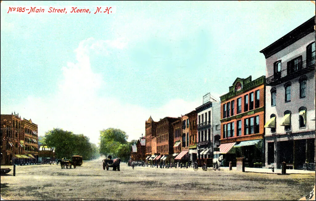 A colored postcard of a wide street bordered by multi-storied buildings. Horse-drawn carriages and wagons can be seen traveling down both sides of the street. The title of the postcard, located in the upper left-hand corner, reads "No. 185 - Main Street, Keene, N.H."