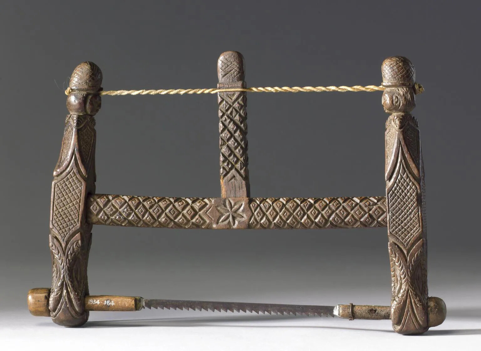 A thin horizontal saw with an ornately carved wooden handle. The handle consists of two vertical pieces of wood, which are connected in the middle by a horizontal piece of wood. A coiled piece of thin rope connects the tops of the vertical pieces of wood with a shorter piece of wood, balanced vertically along the horizontal piece of wood in the middle.