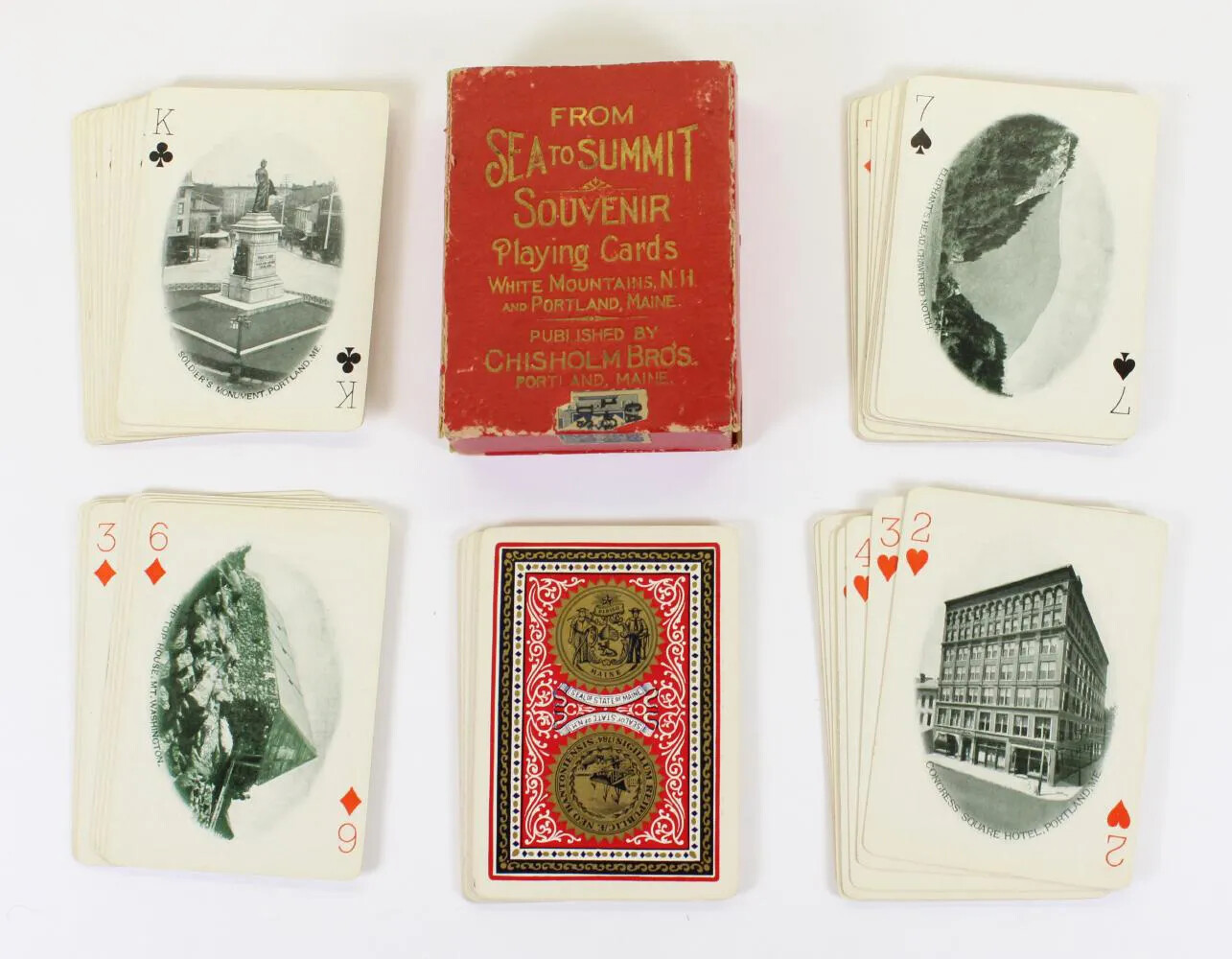 A collection of playing cards, placed in two rows of three piles, with different scenes portrayed in photographs. The card box, placed in the middle of the first row of piles, is red with golden words engraved on top. These words read "FROM/SEA TO SUMMIT/SOUVENIR/PLAYING CARDS/WHITE MOUNTAINS, N. H./AND PORTLAND, MAINE/PUBLISHED BY/CHISHOLM BROS/PORTLAND, MAINE."