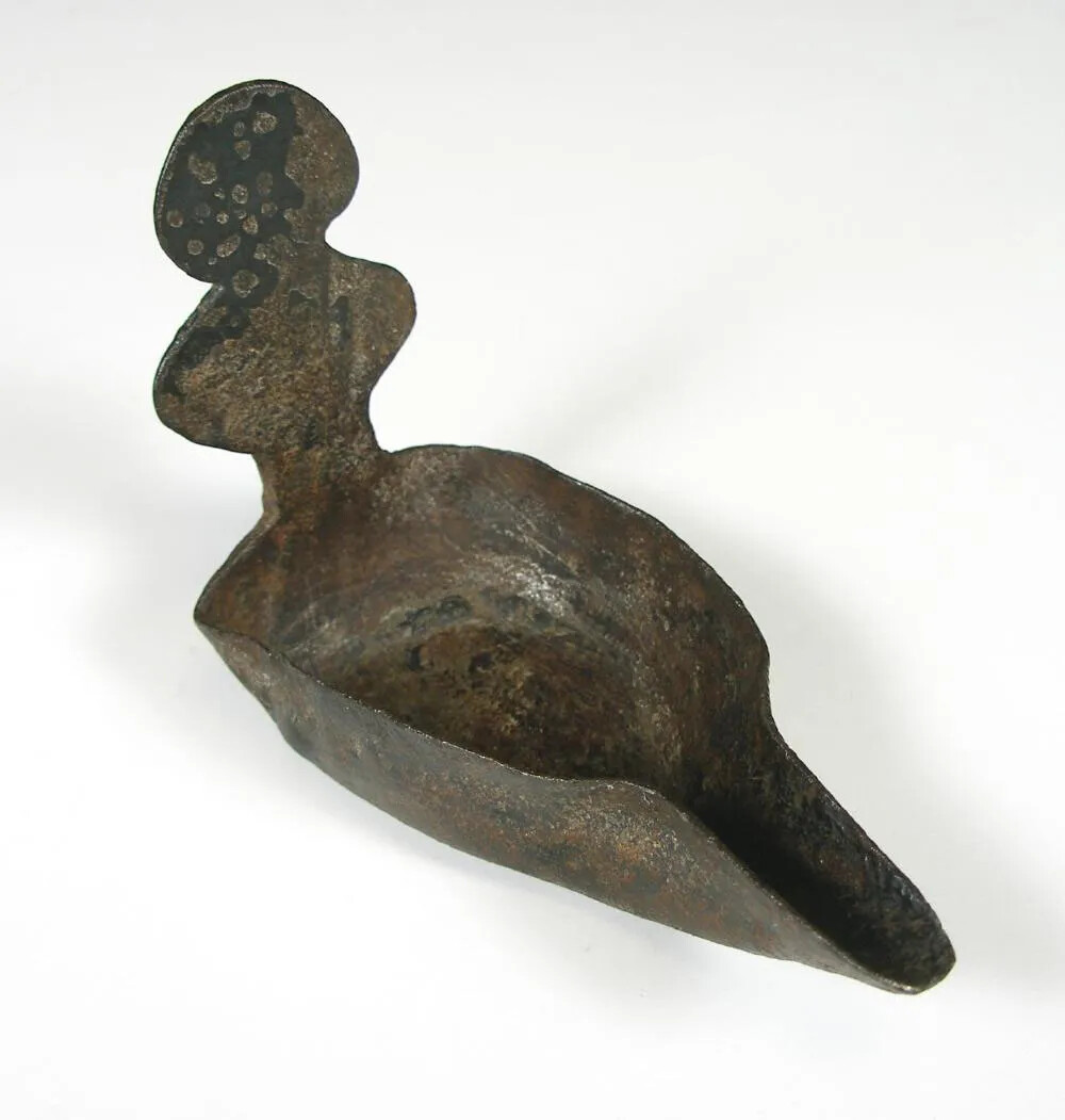 A metal tool with an upright handle. The tool is rounded at the bottom, and has a spouted edge on one side.