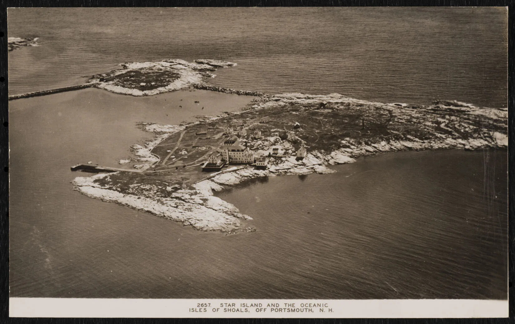 A black and white postcard of two rocky islands, one large and one small. The largest island, placed in the lower half of the postcard, has a few buildings clustered together in the middle of the island. The words "2657 STAR ISLAND AND THE OCENANIC/ISLES OF SHOALS, OFF PORTSMOUTH, N. H." in black text are centered along the bottom border of the postcard.