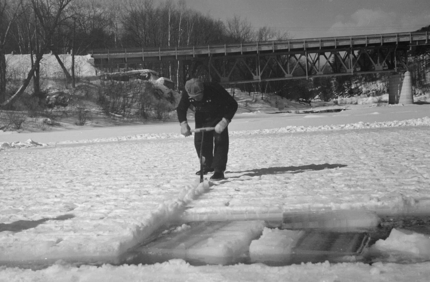 A black and white photograph of a man bent over a thin saw as he cuts through ice on a frozen body of water. In the forefront of the photograph, squares of cut ice float in the exposed water.