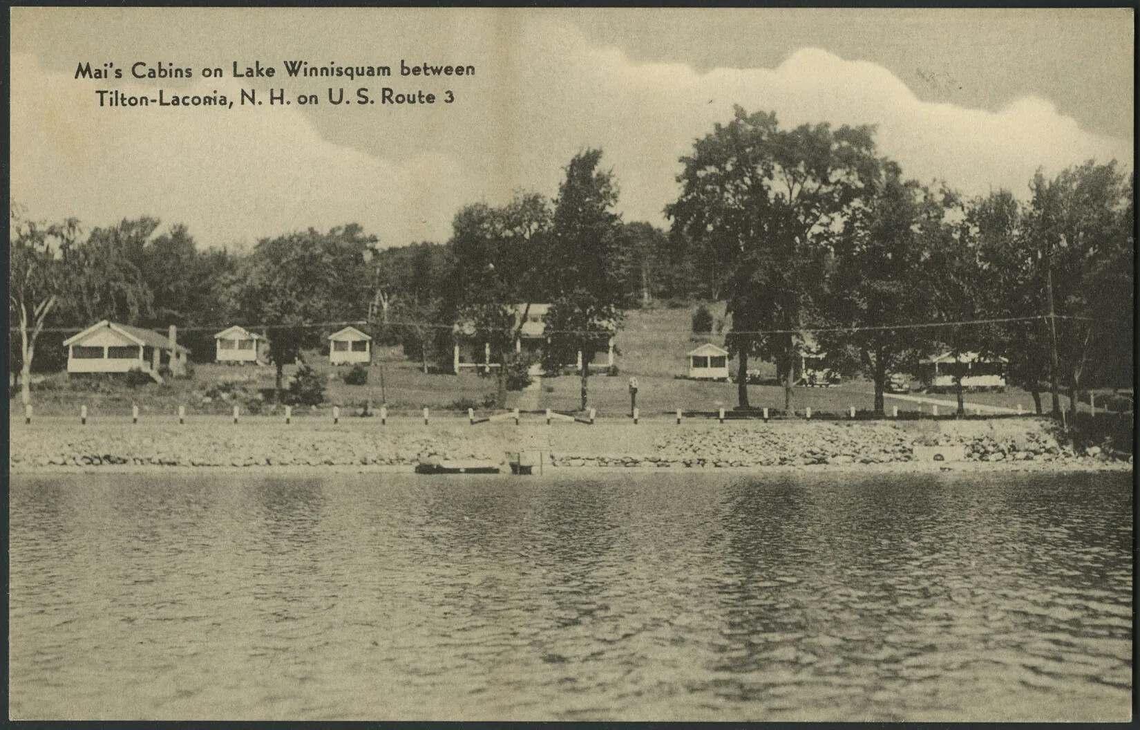 A black and white photograph of a rocky shore and a body of water. There is a small boat in the water at the center of the postcard, and it rests against a small dock. Lining the rocky shore are fence post, and behind this, various single-storied houses are mixed among large trees. In the top left-hand corner, the title reads “Mai’s Cabins on Lake Winnisquam between/Tilton-Laconia, N. H. on U. S. Route 3”.