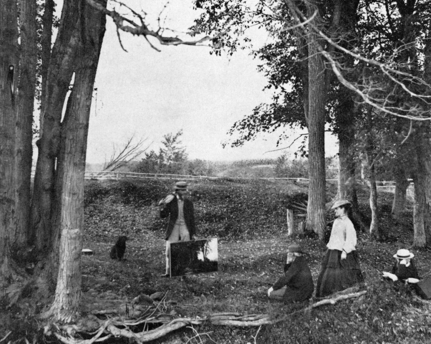 A black and white photograph of a man standing with a painting at his feet in a wooded area. There is a dog seated behind the him. The man faces three people who look at his painting.