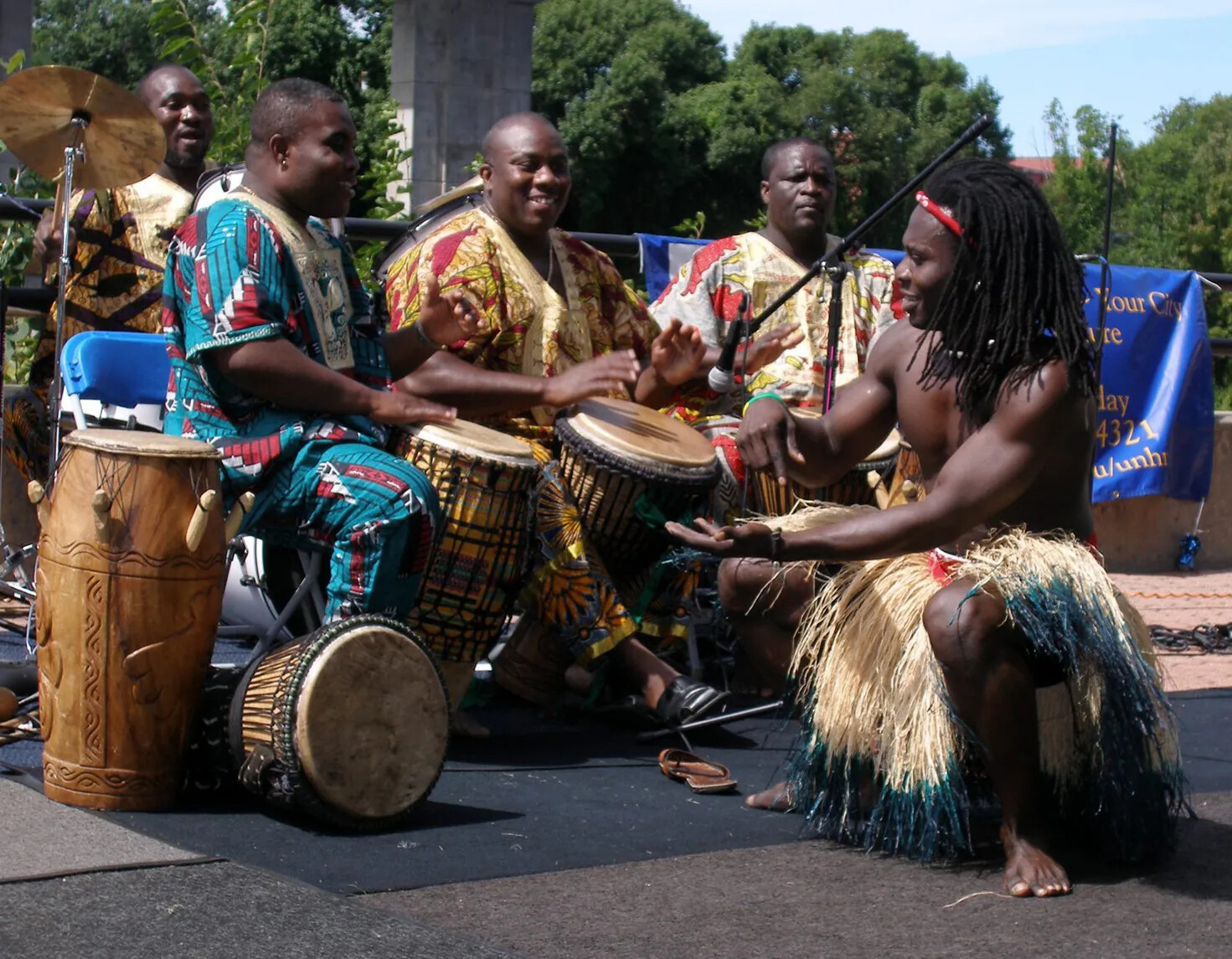 A colored photograph of a group of Black men playing large drums. They all sit in chairs on a stage. One man dances in front of the group of drummers.