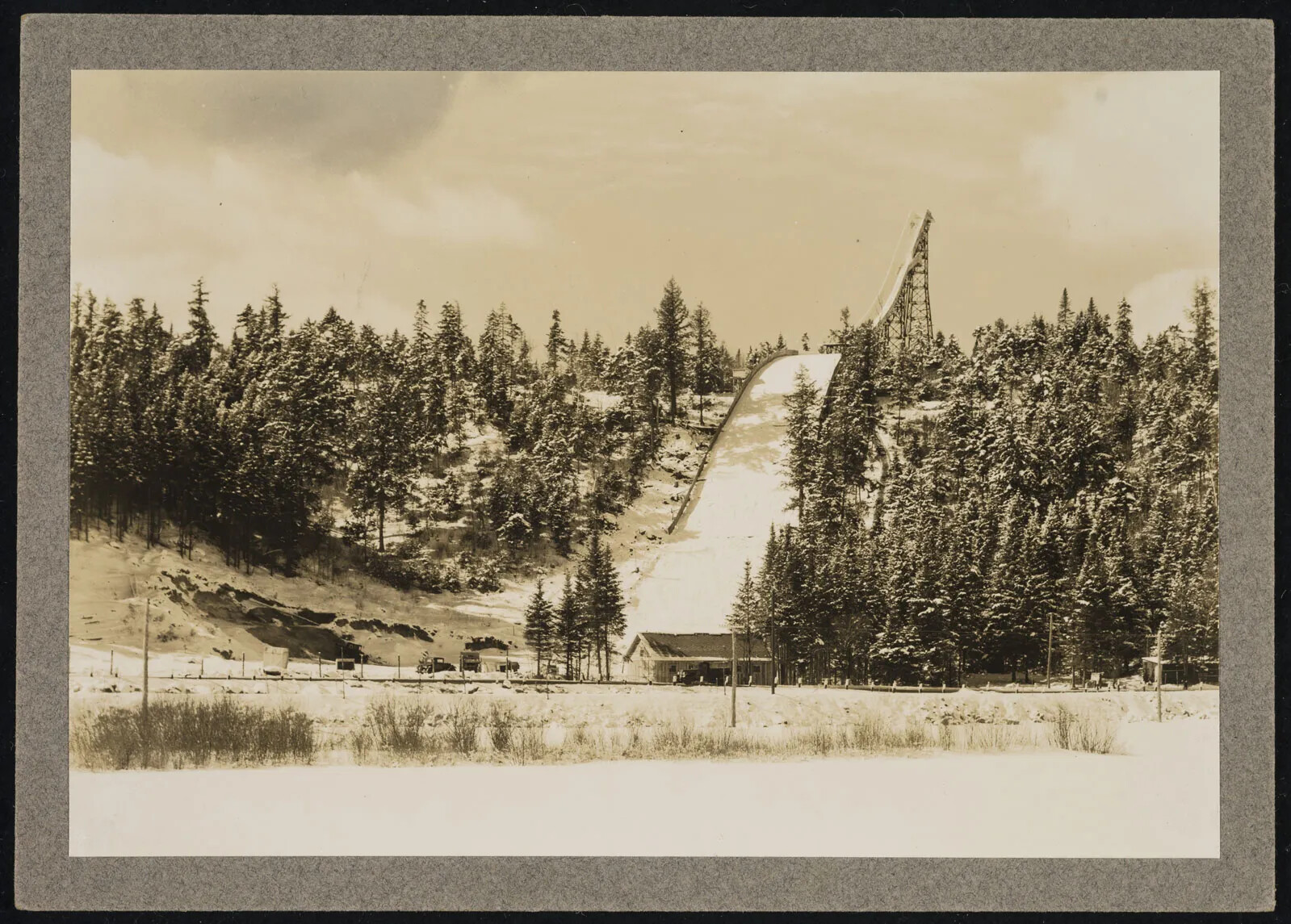 A black and white photograph shows a valley and small hill nearly covered with pine trees in the winter. In the middle of the hill a wide, straight track runs smoothly down the hill, covered with snow. In the distance, a tall tower is built on top of the hill to extend the height of the hill. A narrower, steep track runs down the tower at an angle to meet the track on the hill. A small building sits at the foot of the hill where the track ends. 