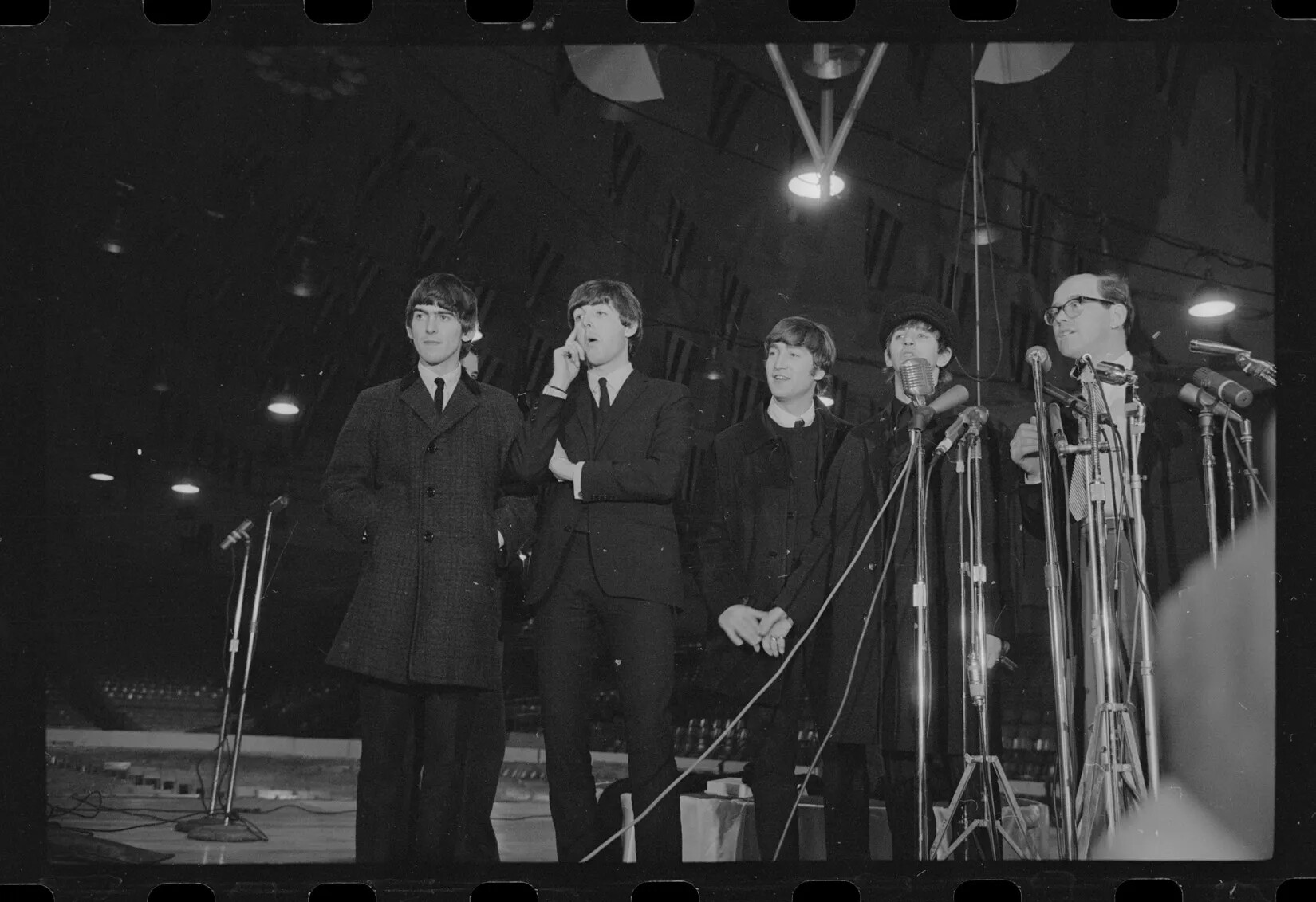 Five men in suits and jackets stand in a line facing the viewer in a black and white photograph. In front of the two rightmost men are many microphones on stands. All the men are looking off to the left, and are standing in a large room with flags hanging from the ceiling. 