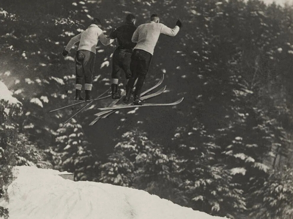 A black and white photograph of three men on skis in mid-air, as if they have just jumped off something. Beneath them is a snowy field, and in the background are snow-covered trees.
