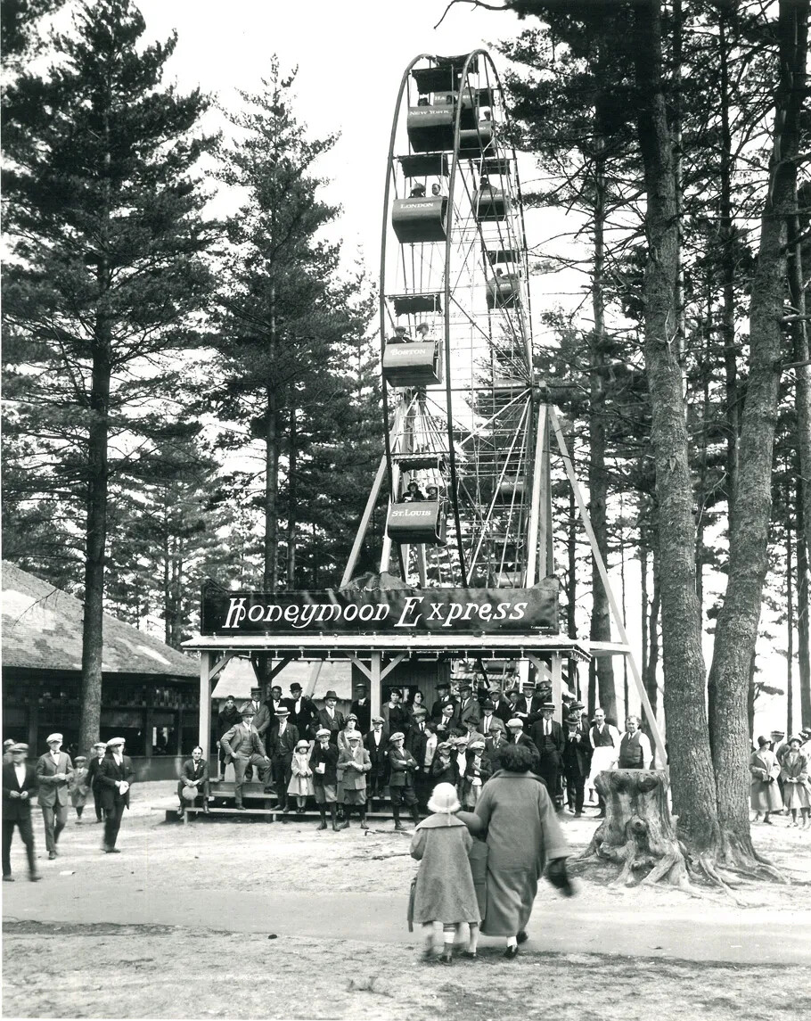 A black and white photograph of a Ferris Wheel in a wooden area. There are groups of people gathered around the base of the Ferris Wheel, with several groups of people walking towards and away from the Ferris Wheel. There is a sign above the entrance to the Ferris Wheel, which reads "Honeymoon Express."