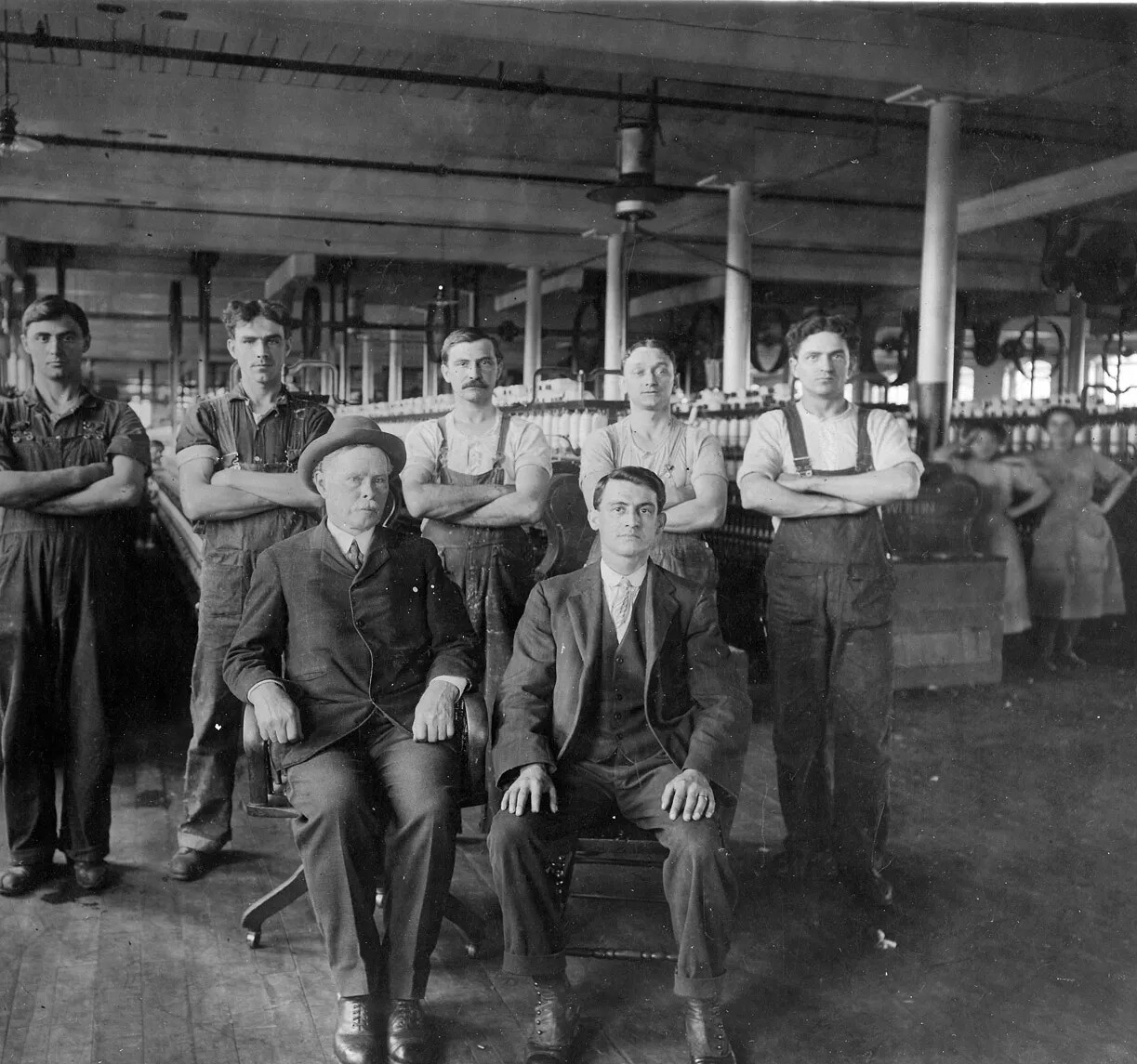 A black and white photograph of a group of seven white men inside a factory. They are posed in front of the camera, with two men in suits seated in chairs, and five men in short-sleeved shirts and overalls standing behind them with their arms crossed. In the background, there are two young women with their hands on their hips and each other who are also starring at the camera.