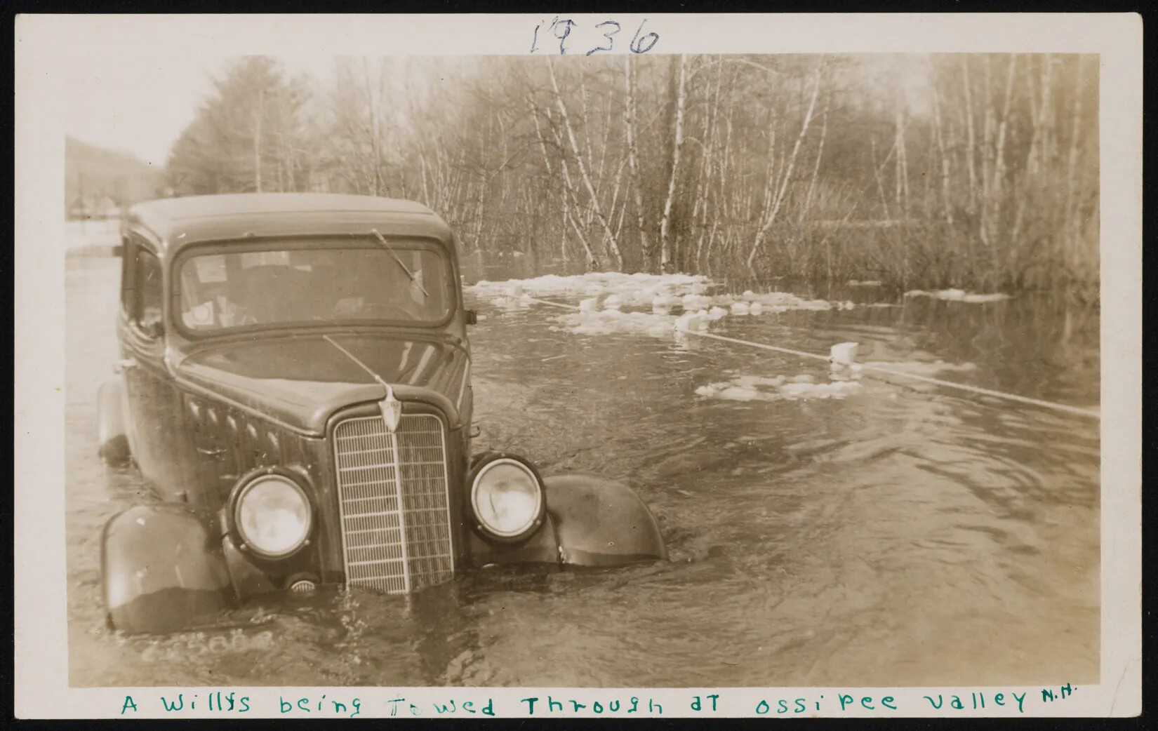 A black and white photograph of a landscape shows a 1930s-style car with flood waters nearly reaching its headlights. Water and some ice flow by the car, and the outline of people are seen inside the car. Flooded birch and other trees are in the background. The top of an almost-covered rail is next to the car. Handwritten penned words read "1936" in the center on top of the photograph, and at the bottom "A Willys being towed through at ossipee valley N.H." 