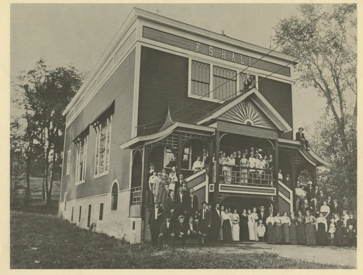 A black and white photograph of a building in a rural area shows many people gathered in the front of the building. They are standing on the porch, stairs, and in front of the building, and are dressed in long skirts and suits. The building is made of brick, has power lines running to it, and has a boxy appearance. At the top of it in large letters reads "F.S. HALL."
