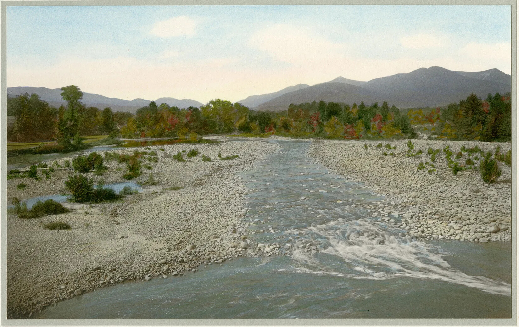 A colored photograph of a waterway passing between rock-laiden land. In the background are trees with green leaves and a mountain range.