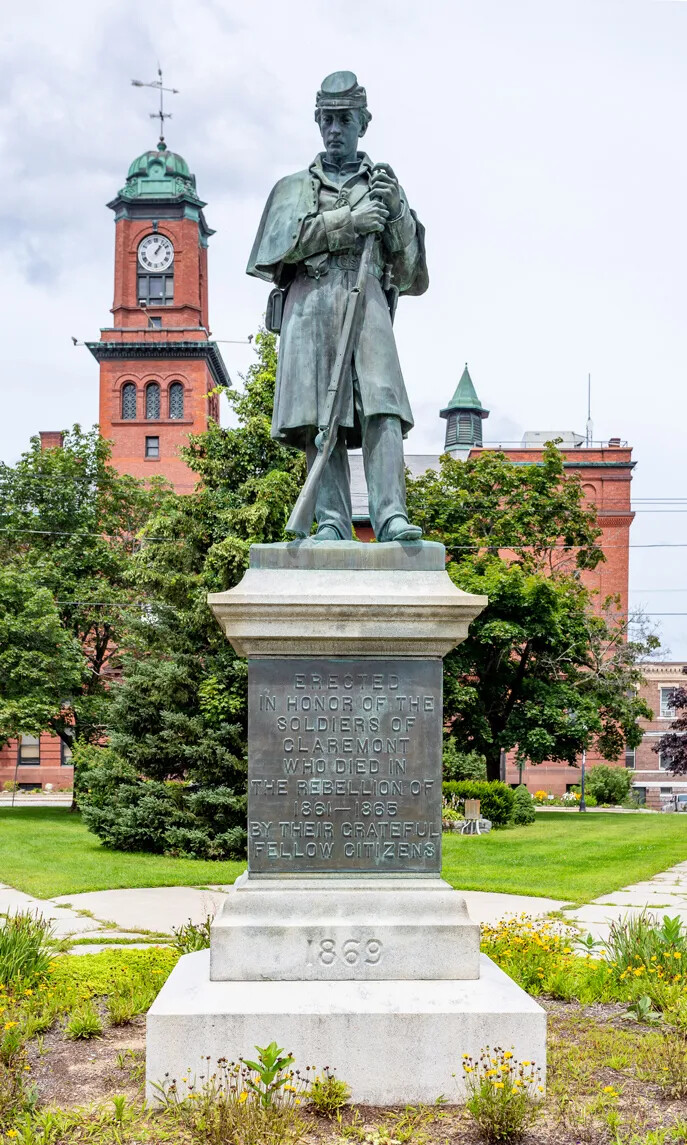 A statue of a man in a flat cap, a long coat, a pair of pants, and shoes. The man looks downwards to the left and holds a musket in his hands. Beneath the man's statue are the words "ERECTED/IN THE HONOR OF THE/SOLDIERS OF/CLAREMONT/WHO DIED IN THE REBELLION OF 1861-1865/BY THEIR GRATEFUL/FELLOW CITIZENS" carved into the pedestal.