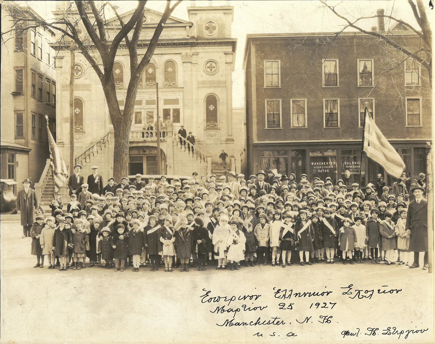 A black and white photograph of a large group of children.  There are some adults standing on either side of the children, as well as on the two-sided staircase in the background of the picture.  Behind the children are three buildings, one of which is a church.  On the left side of the group of children, there is the American flag, and on the right, the Greek flag.  On the right bottom corner of the photograph, there is handwriting in cursive, citing the group’s name, their location, and date of the photograph.