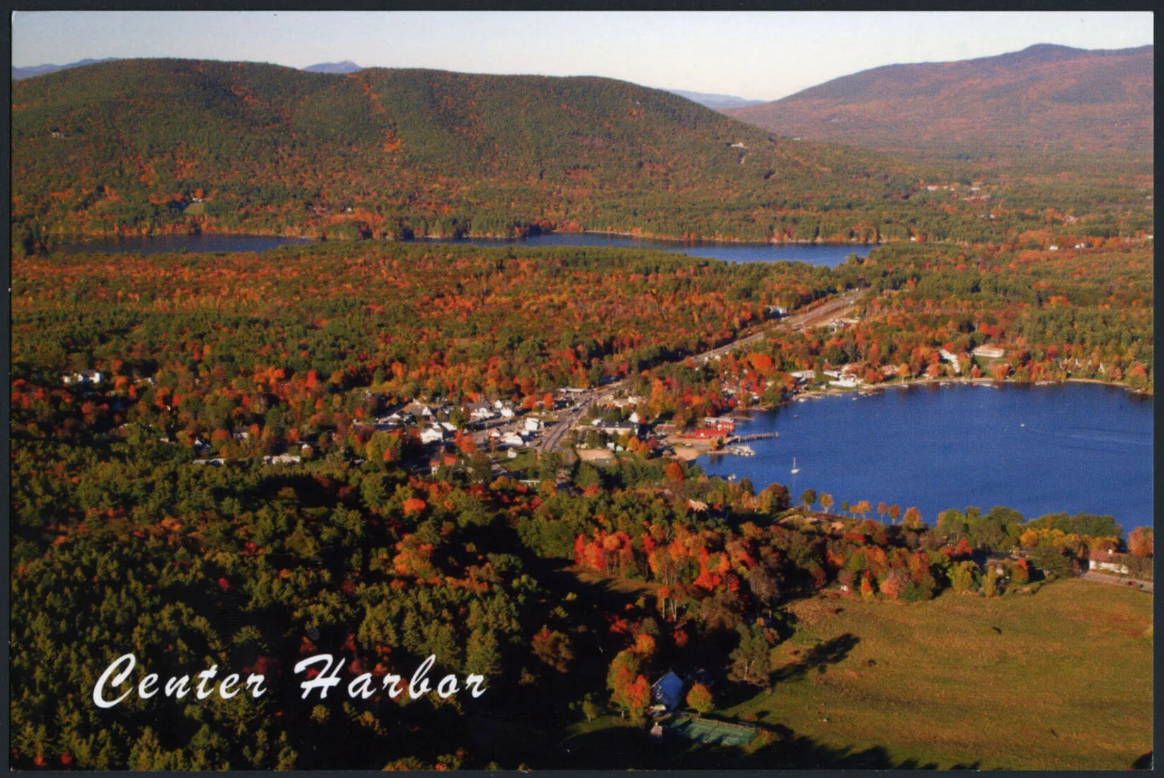 A colored horizontal postcard of a flat area of land covered with trees. There are tall mountains in the background. All of the trees are either green, red, or orange. There is a circular body of water in the right-hand corner, and a another, longer body of water in the upper left-hand corner of the postcard. The title of the postcard, placed in the lower left-hand corner, reads "Center Harbor."