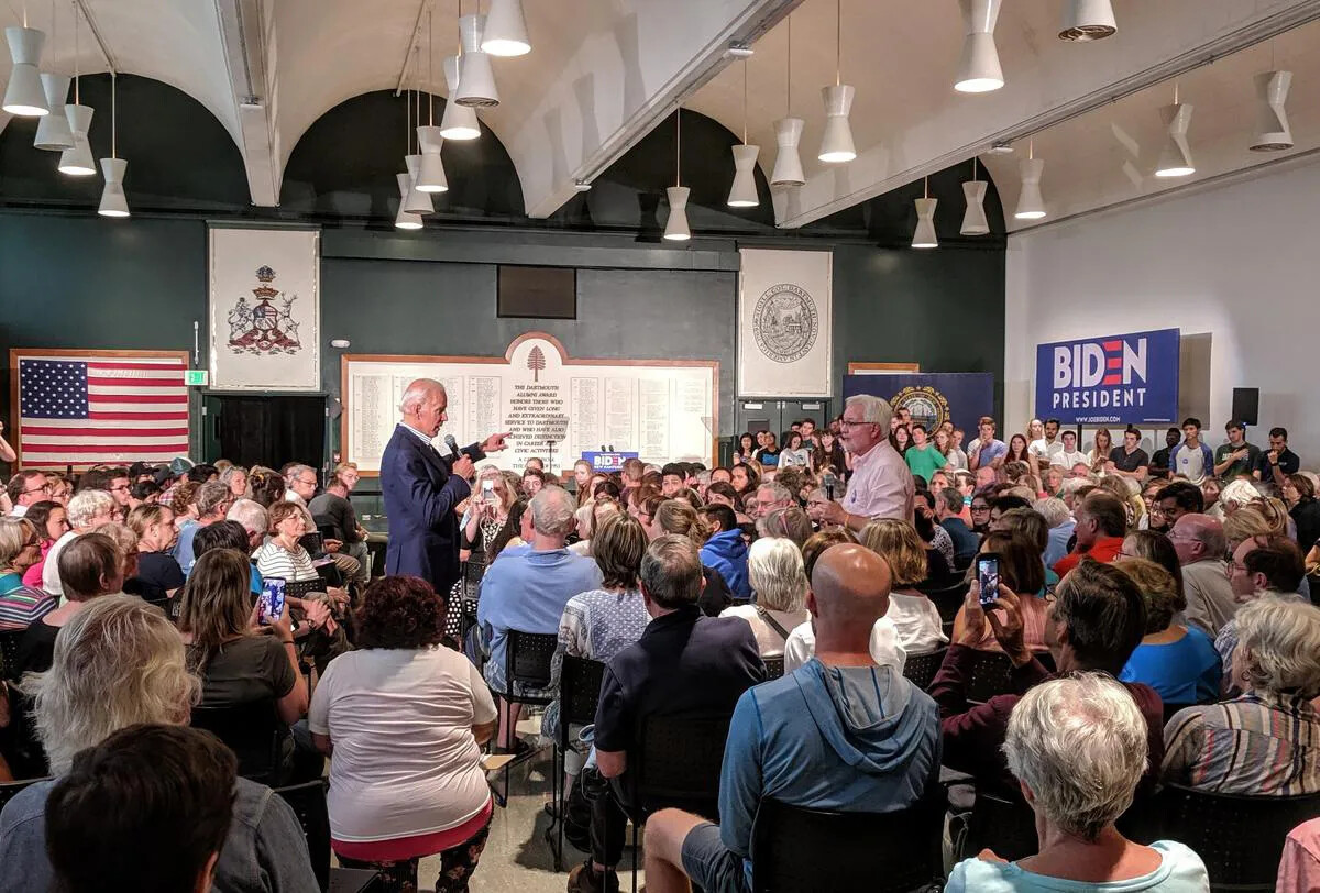 A colored photograph of a man in a blue suit speaking into a microphone. He is standing in the center of a room that is crowded with people seated in chairs.  An American flag hangs from the back wall, and a blue, red, and white sign that reads "BIDEN/PRESIDENT" rests against the right-side wall.