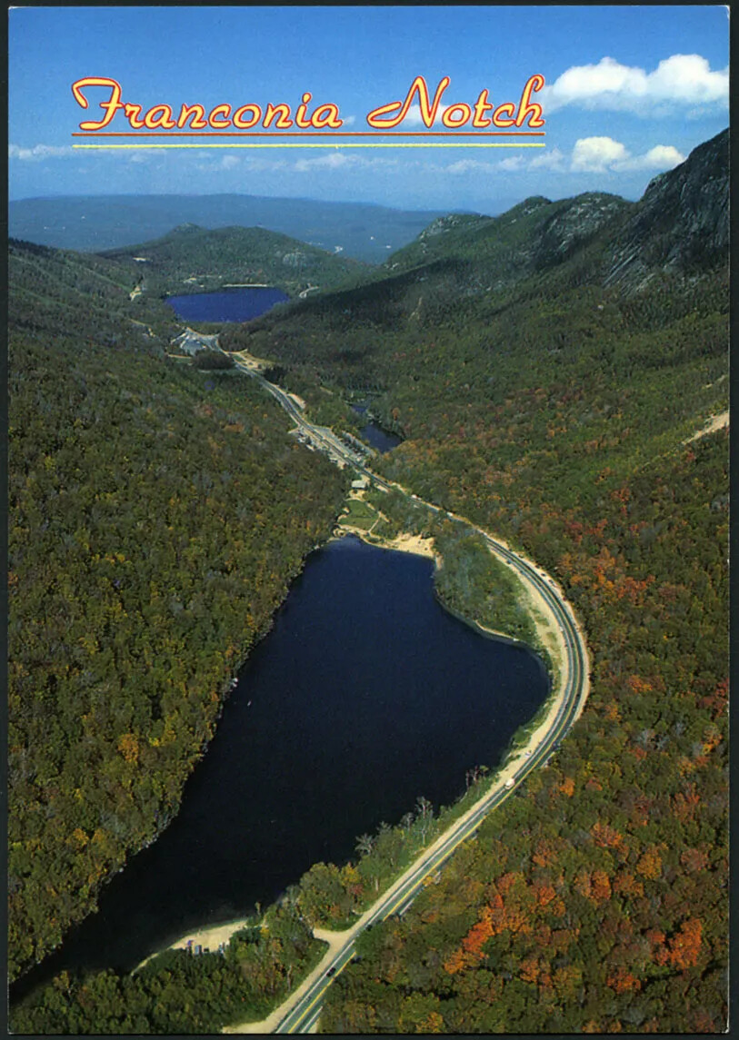 A colored vertical postcard of a body of water surrounded by steep mountains on either side. There is a road to the right of the body of water, and the mountain is covered in green and orange trees. The title of the postcard, placed in the upper left-hand corner, reads "Franconia Notch."