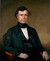 In a color painting, a man sits on a leather chair against a brown background. He is wearing a black suit in the mid-1800s style with high collar and no tie. His hands rest on the arms of the chair in a casual manner, and he looks directly at the viewer with a small smile. He has short curly brown hair and no facial hair. 