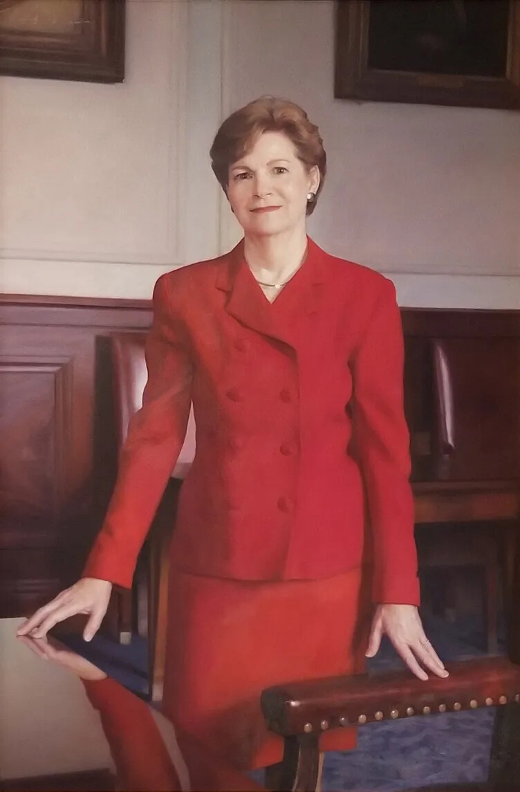 A colored painting of a white woman with short brown hair, dressed in a red jacket and skirt. She stands in front of an armchair and a shiny table.