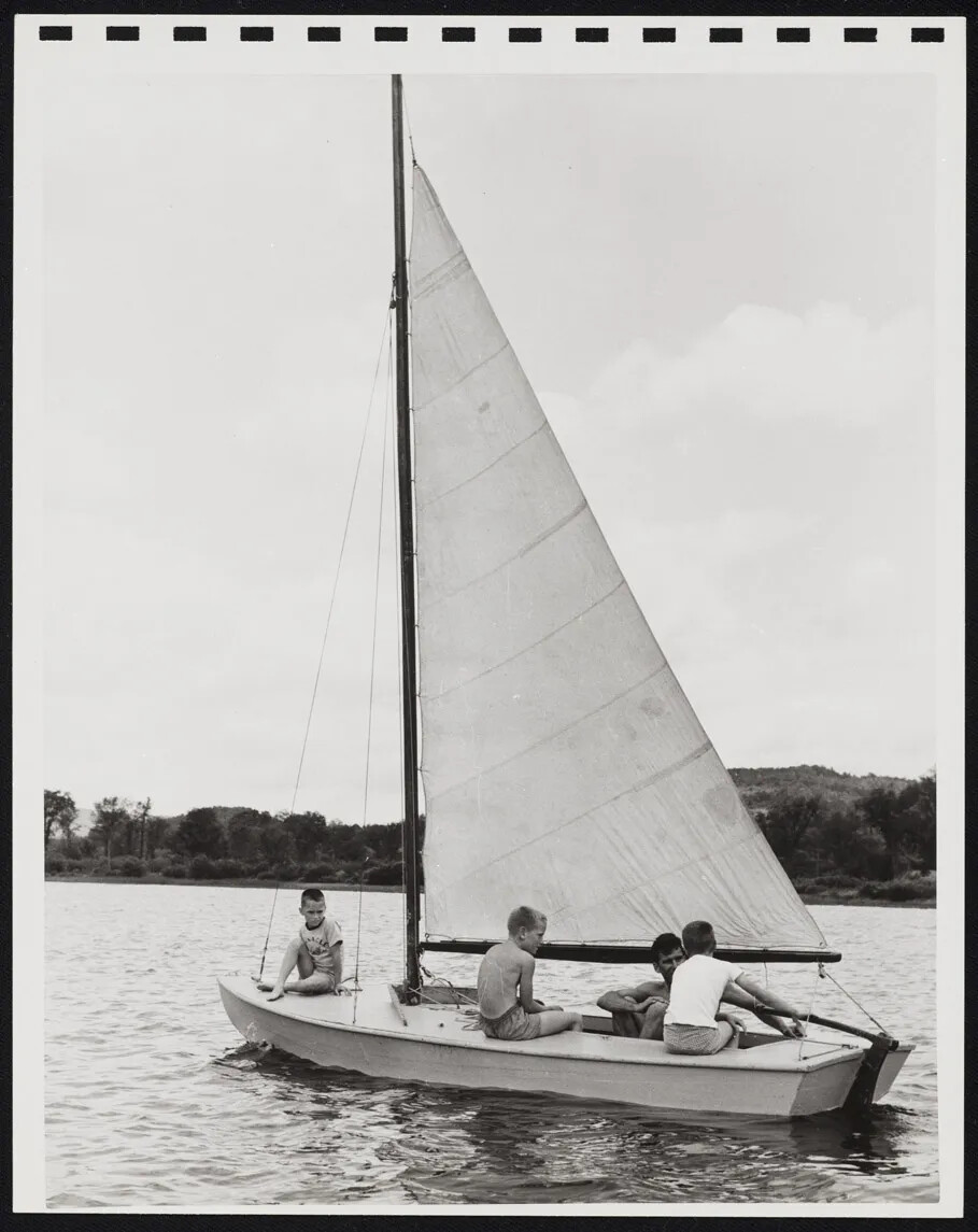 Four figures sail a small sailboat on a lake in a black and white photograph.
