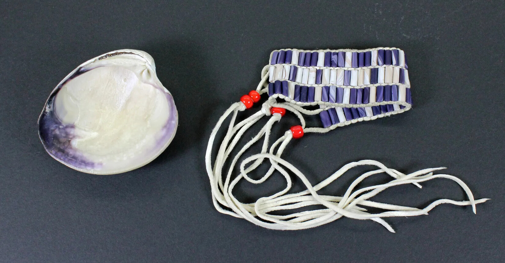 A white shell with purple edges is placed next to a bracelet of alternating white and purple beads. At the ends of the bracelet are long strips of white leather, with red beads knotted at the top of each strip.