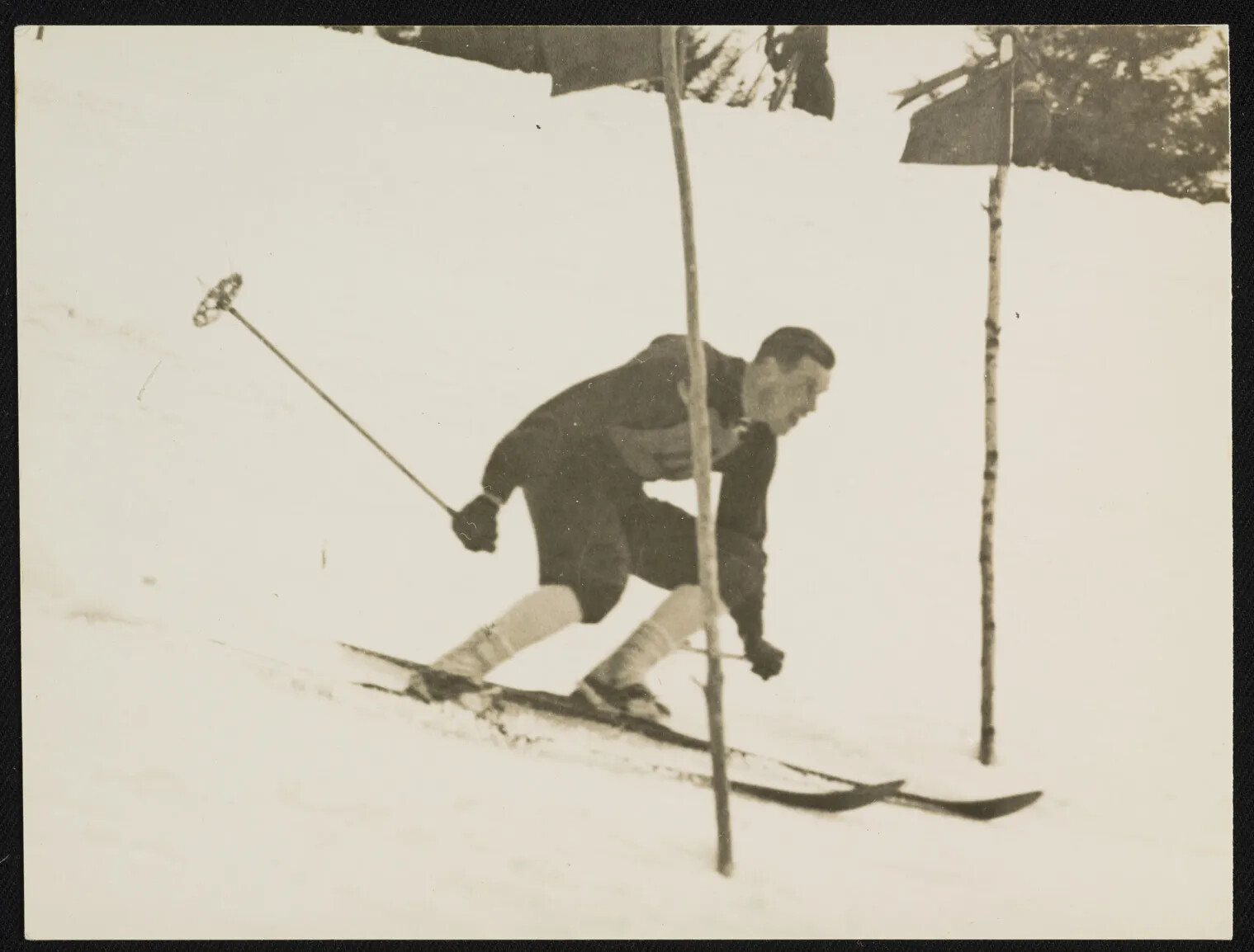 Skier in older sportswear weaves between two birch tree poles as he races downhill in a black and white photograph.