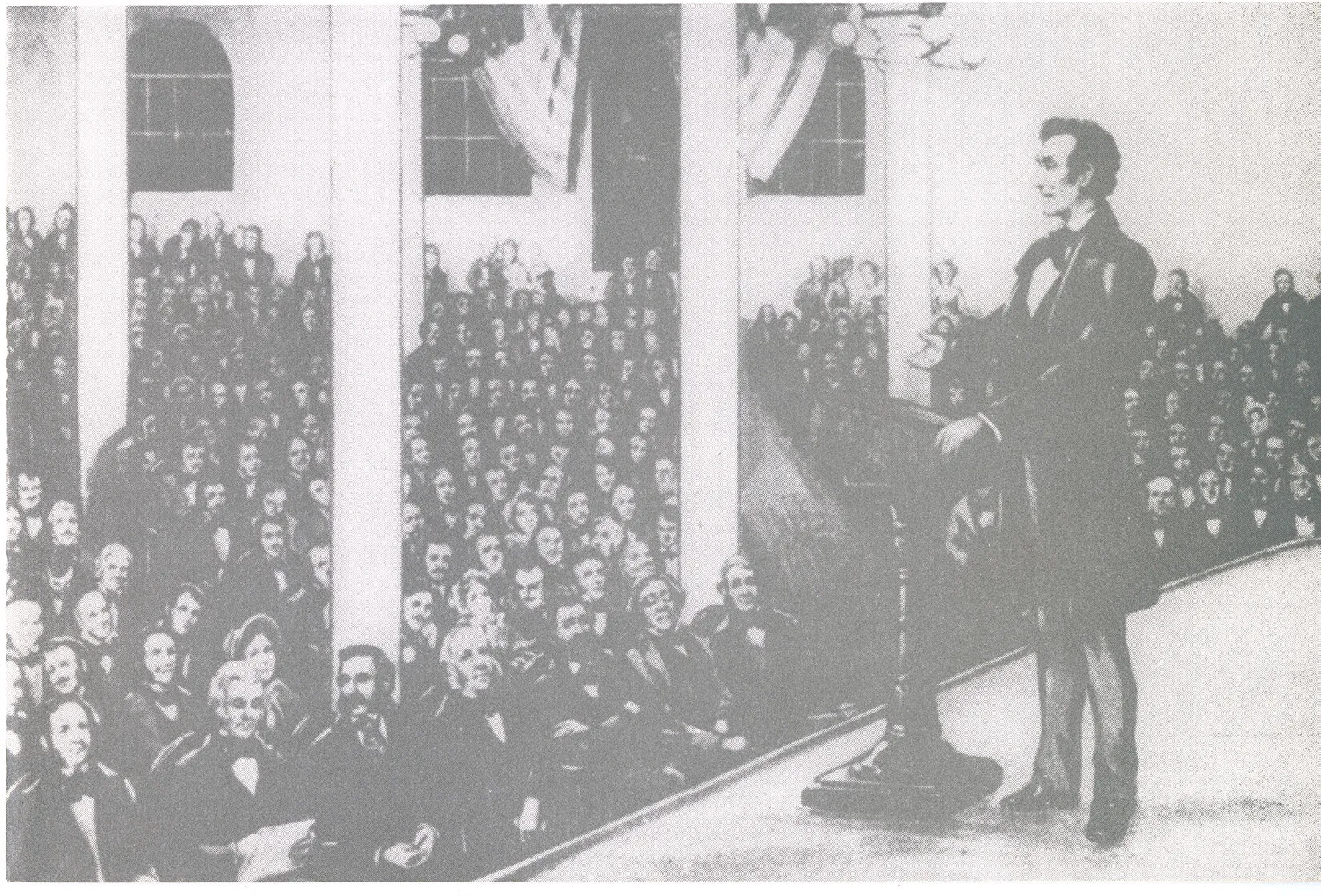 A black and white drawing of a man standing behind a podium on a stage. He speaks to a large group of people seated below the stage.