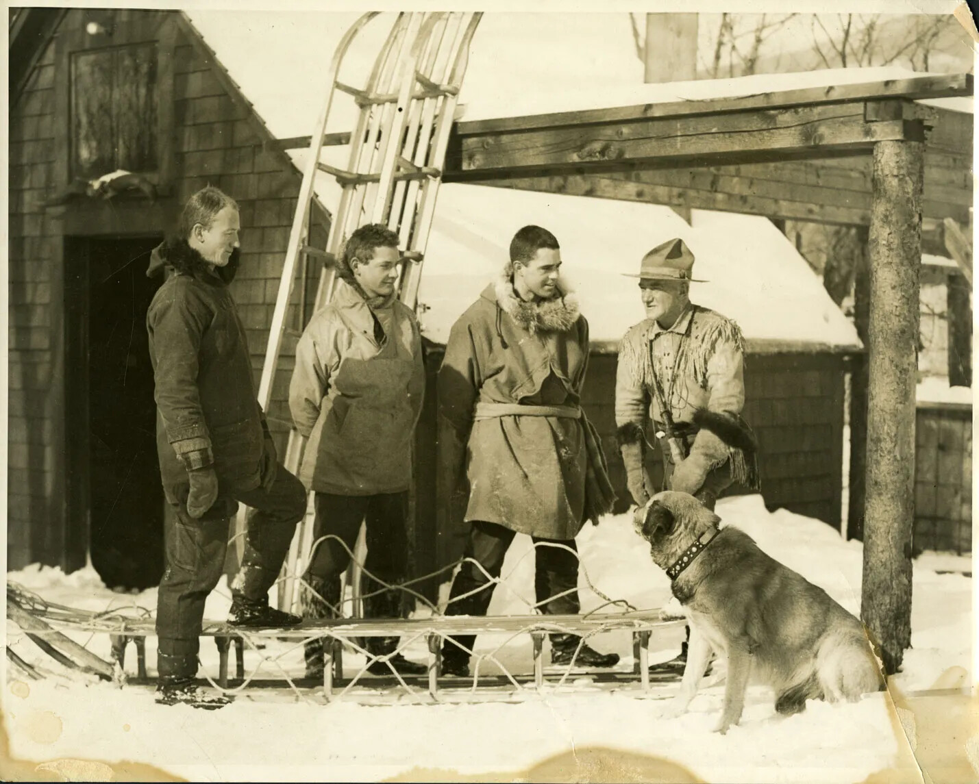 A black and white photograph shows four men and a dog outside. The four men are talking together, and wearing winter outdoors clothes. One man has a hat with a broad rim all around. The dog is sitting looking at the men. There is a barn and lean-to in the background as well as two toboggins. One toboggin is lying in front of the men and one man has his foot on it; the other toboggin is standing up against the lean-to behind them. 