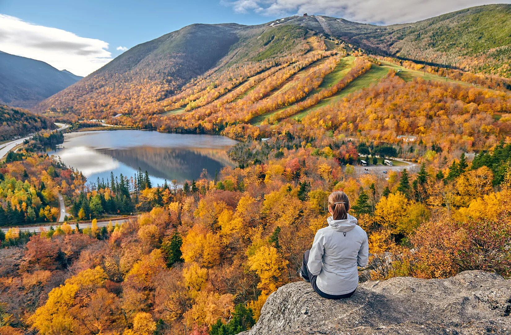 A colored photograph of a woman with her back to the viewer, sitting on the edge of a cliff. She overlooks a valley with a lake and mountain range filled with multi-colored trees.