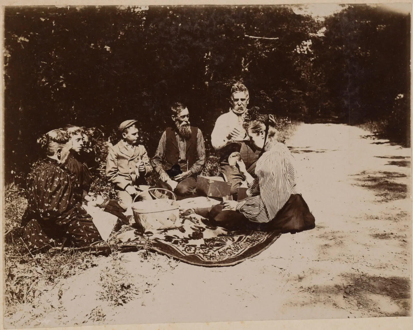 A black and white photograph of a family of six having a picnic on the side of a road in a forested area. The family, from left to right, has two women in long dresses; a boy in a collared shirt, a jacket, and a cap; two men with beards and mustaches in pants and long-sleeved shirts; and a women in a long skirt, a blouse, and a bonnet. They all sit in a semi-circle on a floral carpet.