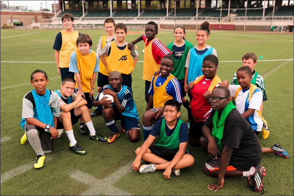 A colored photograph of a group of children of multiple races, wearing soccer gear. They stand and sit on a green field with white lines drawn on it. In the background are rows of seats placed in an upward angle.