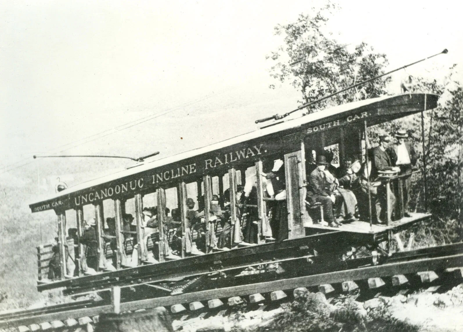 A black and white photograph of a trolley filled with people, traveling up a steep mountain side on a set of railroad tracks. Painted onto the side of the trolley are the words "UNCANOONUC INCLINE RAILWAY."
