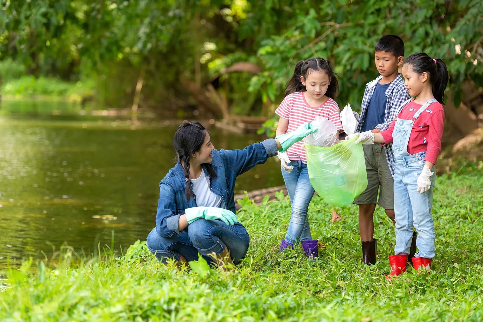 A colored photograph of a woman and three children cleaning up garbage on a grassy area next to a body of water.