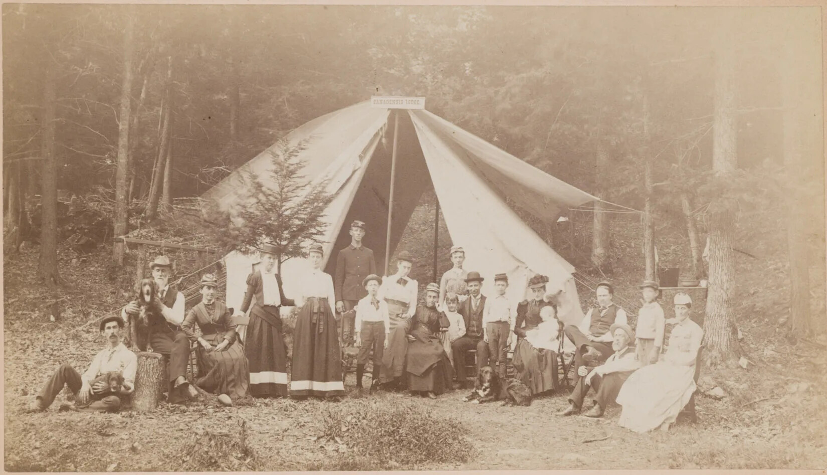 A black and white photograph of a large group of men, women, and children sitting and standing in front of a large circular tent. There are three dogs scattered among the group as well. The group poses facing the camera in a heavily forested area. The tent is held up by poles and by numerous ropes tied to the surrounding trees.