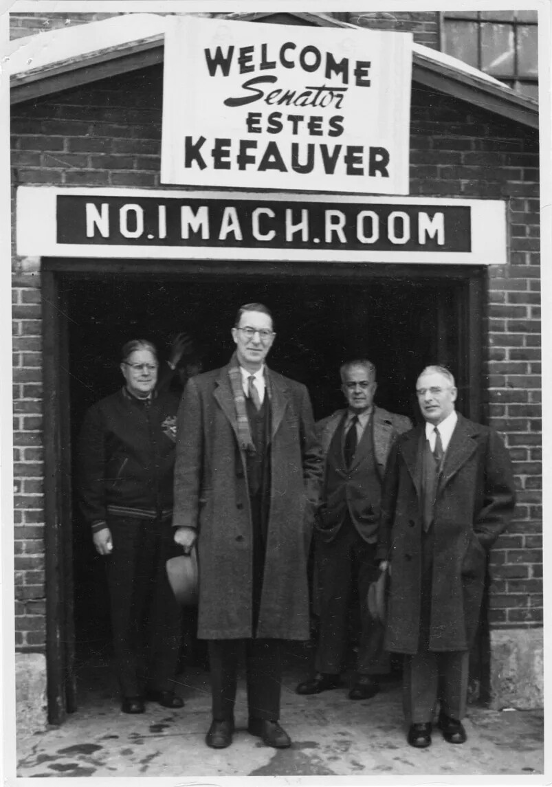 A black and white photograph of four men standing in the doorway of a brick building. Two men stand slightly in front of the other two. The men in front carry their hats, and all are wearing winter clothes. A sign placed above the front door reads "WELCOME/Senator/ESTES/KEFAUVER." Directly below this welcome sign is another sign, which reads "NO. 1 MACH.ROOM."
