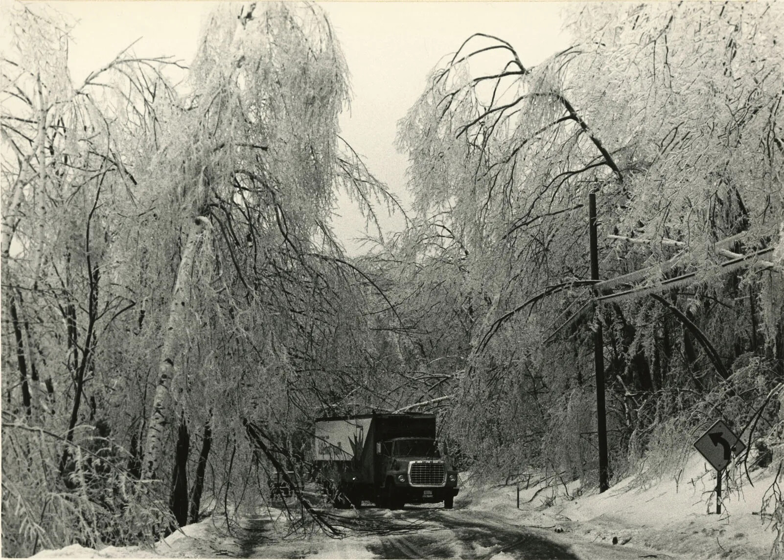 A black and white photograph of a truck driving along a road lined with ice-covered trees.