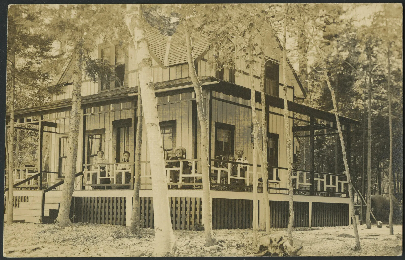 A black and white postcard of a two-story, gabled house surrounded by a forested backdrop and individual trees scattered around its front and side. The house is decorated with wood paneling and has a wrap-around porch with a railing. Sitting on the porch, from left to right, are two women, a man, and a man with a small child. Everyone on the porch pose looking toward the camera.