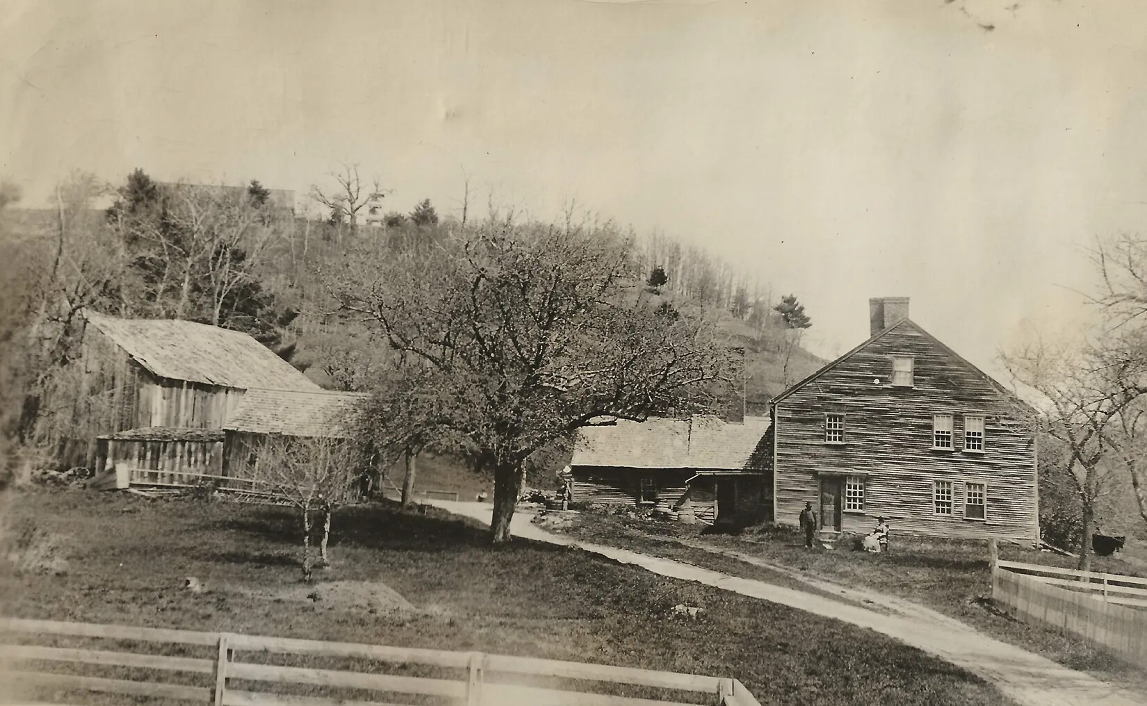 A black and white photograph of a farm with several buildings, including an L-shaped house and a barn. The front part of the house is two-storied. The boundaries of the farm yard are fenced off with horizontal wooden posts, and the land is scattered with trees.
