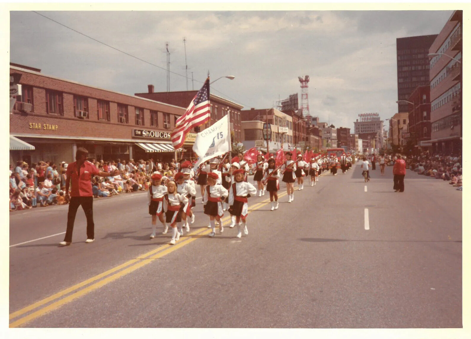 A colored photograph of a group of children marching in a parade. They all wear red caps, white shirts, black shorts, and white sneakers. One of the children holds a large American flag, and another holds a smaller white flag with some writing on it. The road they are walking on is lined with two- and three-storied buldings and people sitting and standing on the sidewalks.