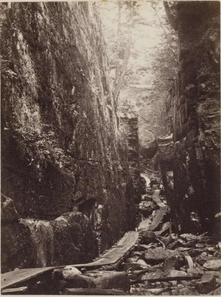 A black and white photograph of a narrow, wooden foot bridge, placed over a rocky floor, with two walls of rock on either side. 