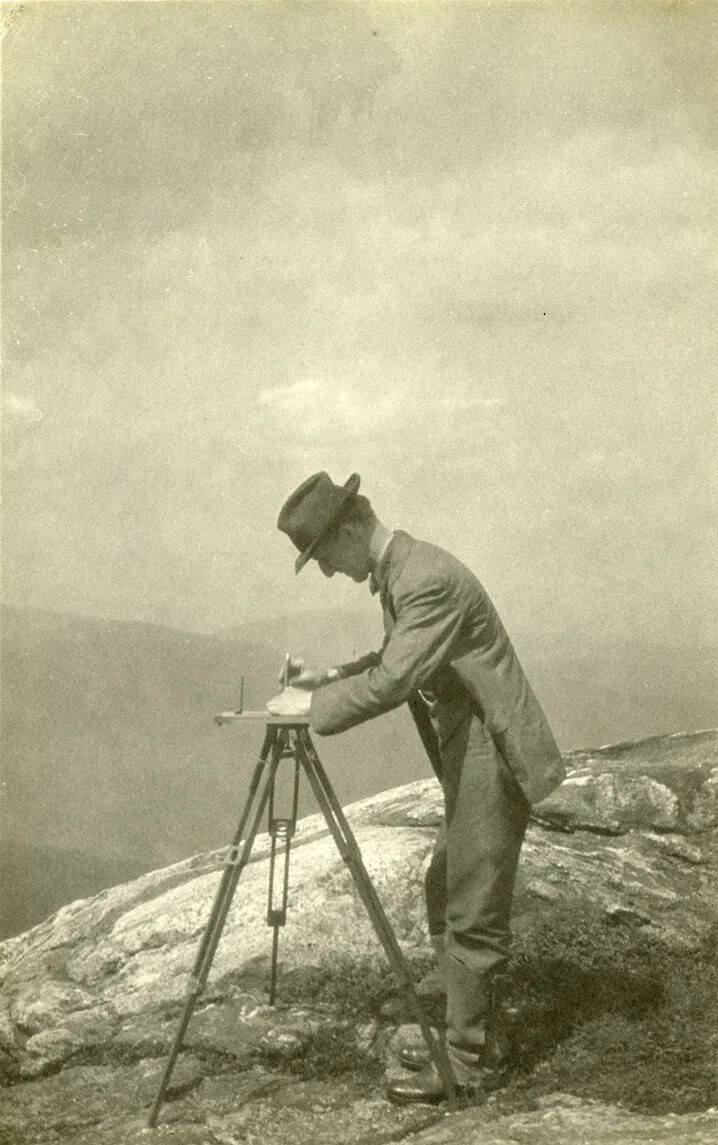 A black and white photograph of a man in a suit and hat using a tripod on top of a mountain. In the background are the outlines of other mountains in the area.