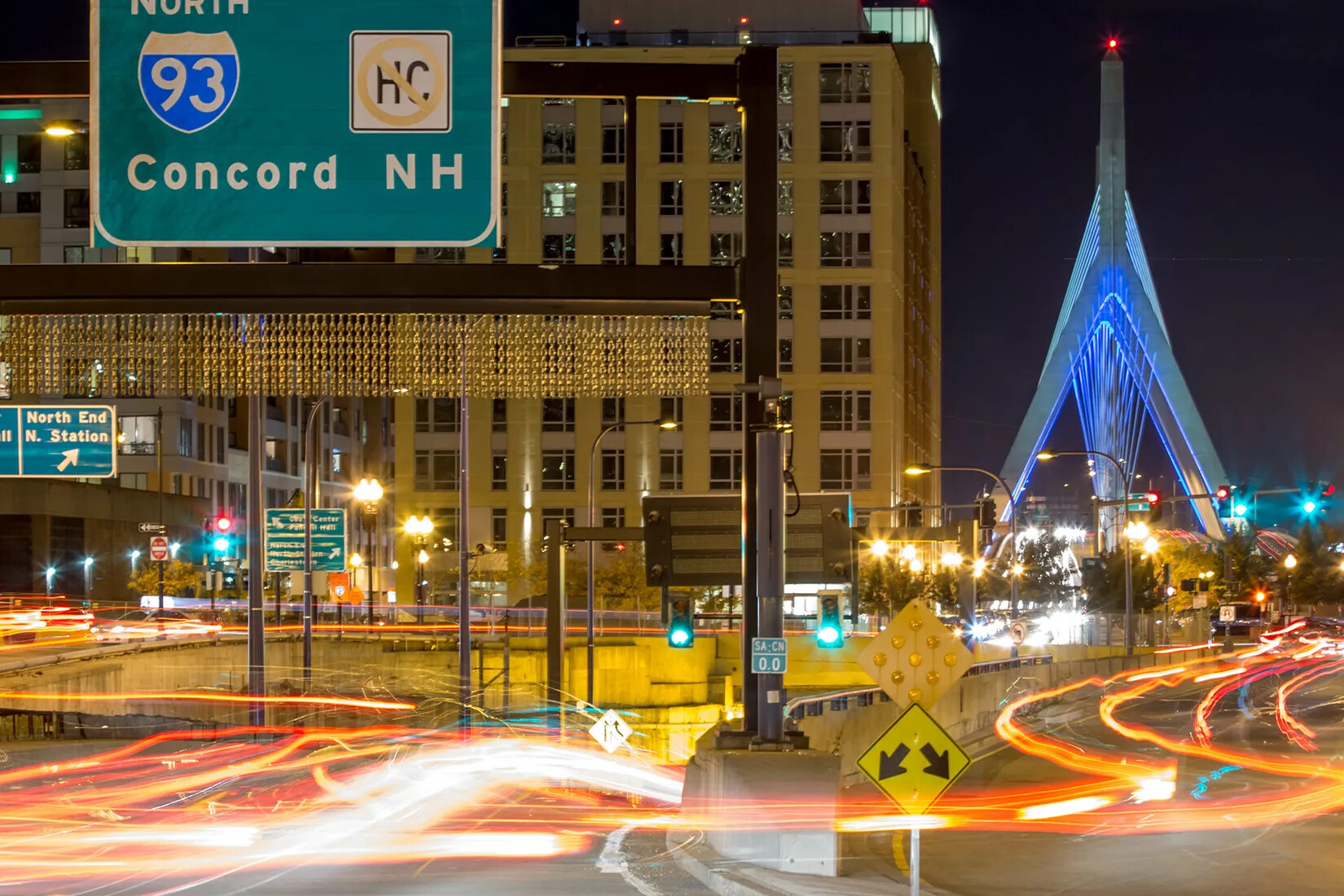 A colored photograph of a road with multiple on-ramps. Blurred lines of red and white travel along the road. In the background is a multi-storied yellow building and a triangular bridge. Above the road is a green sign with the words "Concord NH" typed in white text.