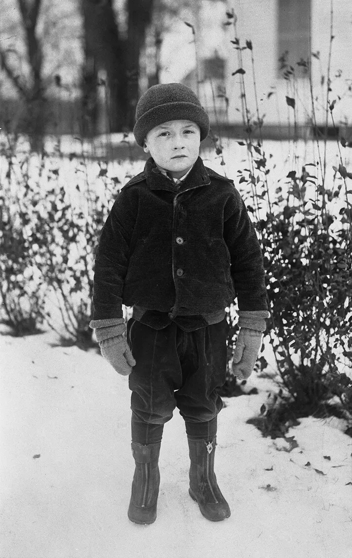 A black and white photograph of a little boy standing outside in the snow. He looks directly at the viewer. He wears a hat, coat, gloves, pants, and tall boots.