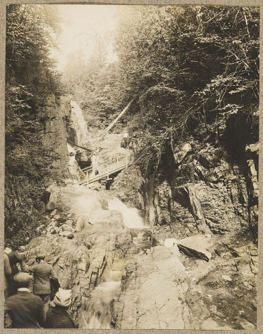 Black and white photographic print of the a waterfall cascading through a small gorge with many rocks and trees around it. There is a a bridge at the center, and a group of unidentified individuals in the lower left corner.